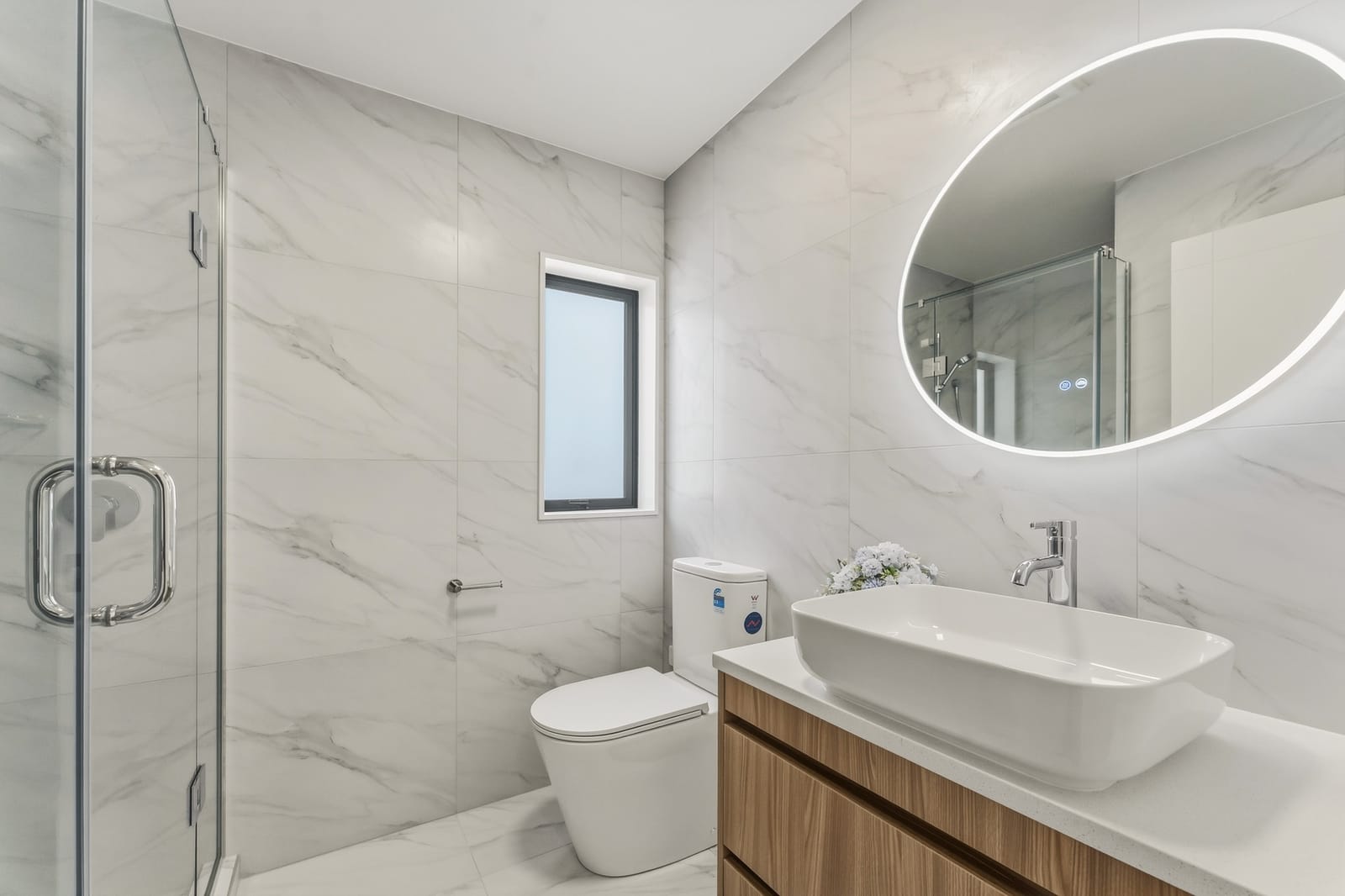 Bathroom with a glass shower, backlit oval mirror and marble-look tiles
