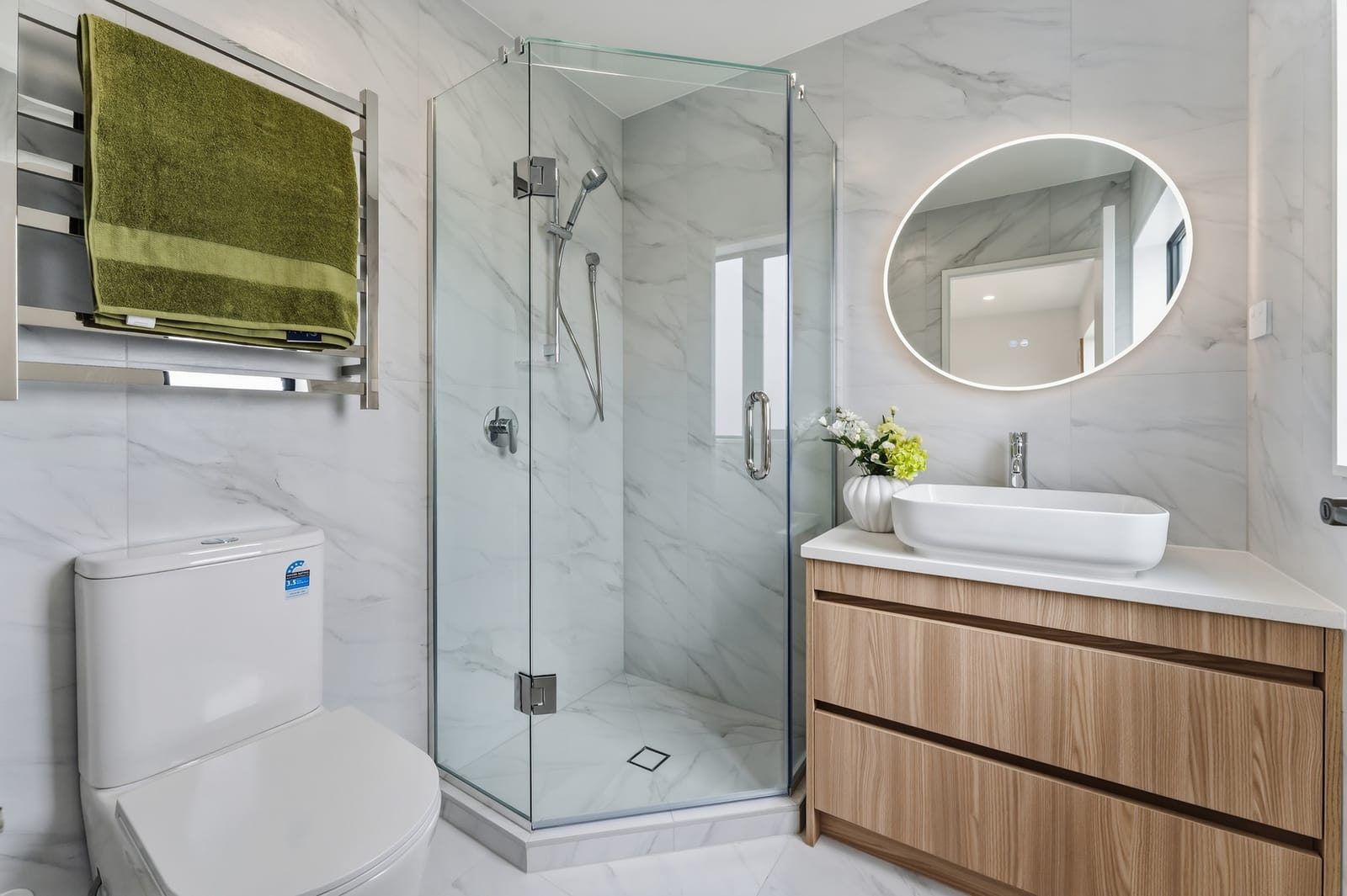 Bathroom with a chrome towel ladder, corner shower and backlit oval mirror