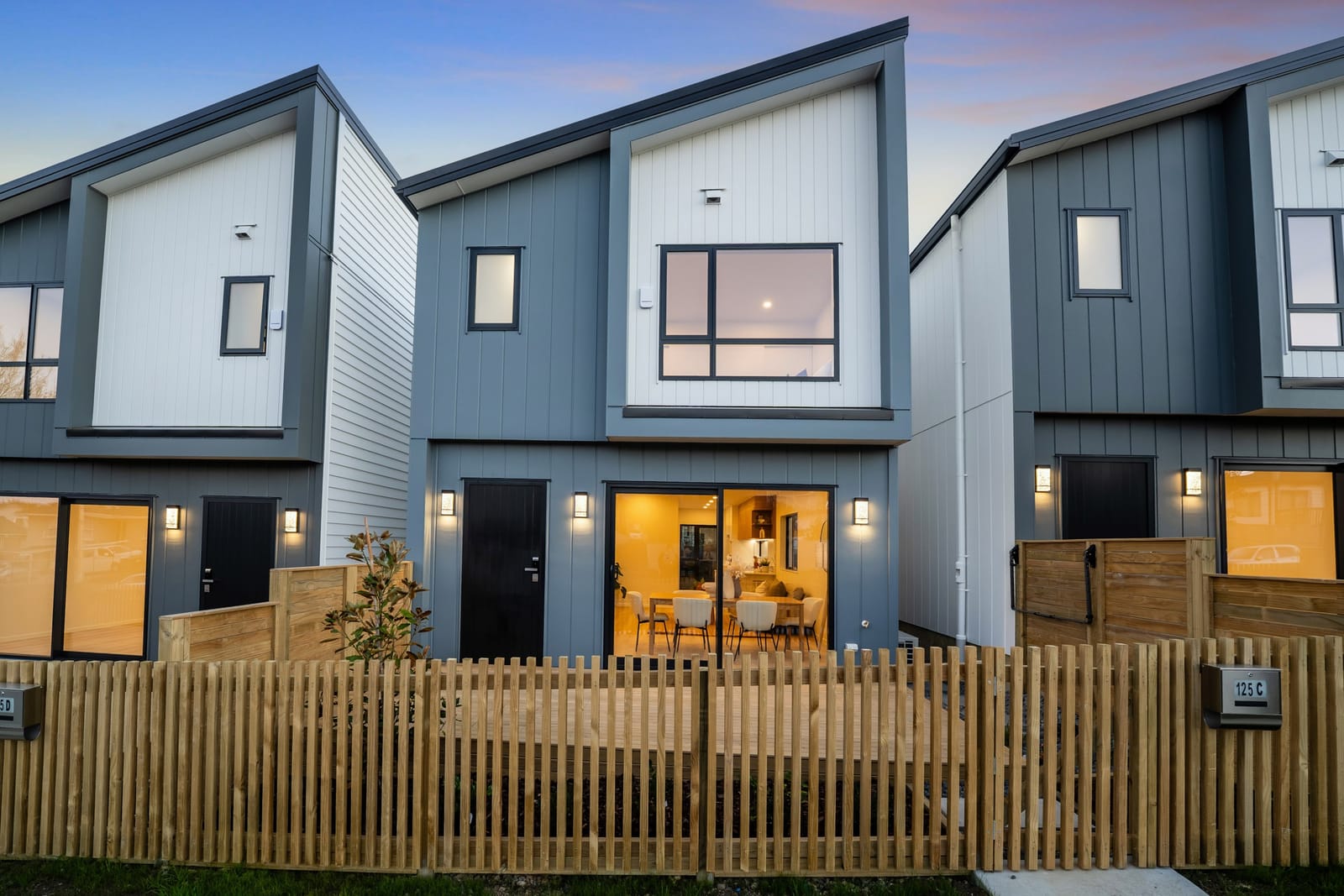 The fronts of the homes at twilight, warm light from sliding doors falling on private decks behind a timber fence