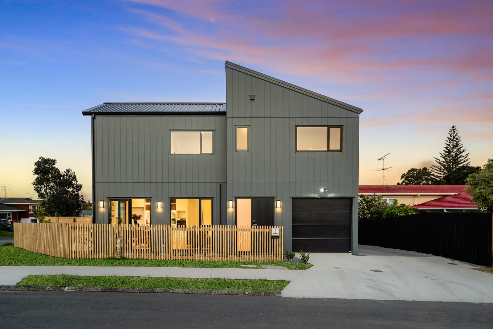 Lot 1 at dusk: a two-storey home in dark grey vertical cladding with a skillion roof, lit windows, a garage and a slatted timber fence