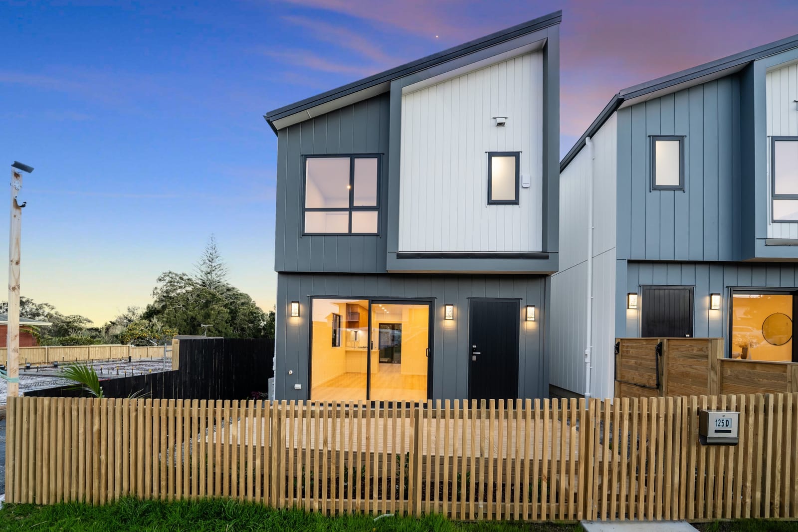 Lot 5, the end home, at twilight, in white and grey cladding with its living area lit behind a timber fence